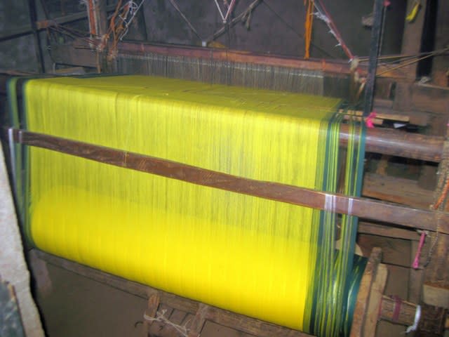 A warp beam loaded on the handloom with warp held in tension. Also, warp ends passing through eyes holes of the heddles may be observed.